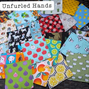 Op de afbeelding: Een verzameling kleurrijke, vierkante stoffen items met verschillende patronen, waaronder appels, citroenen, honden, katten en schapen. De tekst "Unfurled Hands" staat bovenaan. De items zijn in een overlappende manier gerangschikt.