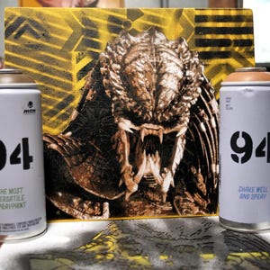 Puede incluir: Una obra de arte de Predator sobre un panel de madera, con un fondo geom&eacute;trico amarillo y negro. Varias latas de pintura en aerosol con el n&uacute;mero "94" est&aacute;n frente a la obra de arte. Las latas son de color lila claro con tapas marrones.