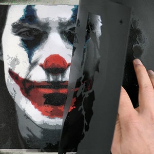 May include: A canvas painting of the Joker with red, white, and blue accents. A black stencil is being lifted to reveal the artwork. Spray paint cans are visible in the background, suggesting an art project.