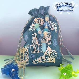 May include: A teal drawstring pouch with white printed designs and the text "Skylanders Trap Storage Pouch". A beaded necklace with a blue crystal pendant hangs from the pouch. Several translucent toy figures are in the foreground.
