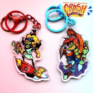 May include: Two clear acrylic keychains featuring characters from the Crash Bandicoot video game series. The keychain on the left features Dr. Neo Cortex, a mad scientist, riding a rocket. The keychain on the right features Crash Bandicoot, a bandicoot, holding a gem and riding a rocket. Both keychains have blue and red key rings.
