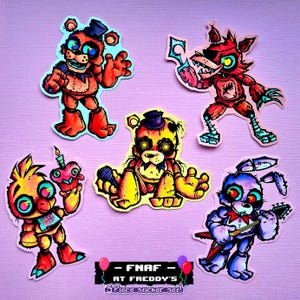 Puede incluir: Juego de cinco pegatinas con personajes de dibujos animados de la serie de videojuegos Five Nights at Freddy's. Las pegatinas incluyen a Freddy, Foxy, Chica, Golden Freddy y Bonnie, con el texto "FNAF AT FREDDY'S".