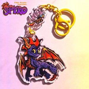 May include: Clear acrylic keychain featuring a purple dragon with orange wings and horns, and a small insect. The keychain has a yellow chain and clasp. The text "Spyro" is visible in the upper left corner.