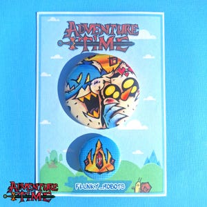 May include: Two Adventure Time pin badges on a light blue card. The top badge features a colourful, expressive character design. The bottom badge displays a gold crown design. The card has the Adventure Time logo and "FLUNKY_ROBOTS" text.