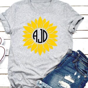 May include: A gray t-shirt with a yellow sunflower design. The sunflower has a black circle in the center with the letters "AJD" inside.