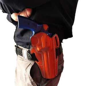 Peut inclure: Un étui en cuir marron avec une arme bleue à l'intérieur. L'étui est porté sur la hanche d'une personne et fixé par une ceinture.