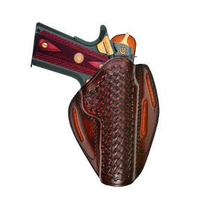 Könnte beinhalten: Ein braunes Lederholster mit einem gewebten Muster und einer Pistole darin. Die Pistole hat einen rot-schwarzen Griff und eine goldene Verzierung.