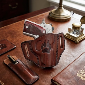 May include: A brown leather holster featuring an embossed paw print design, containing a silver and wood-gripped semi-automatic pistol. The holster includes belt slots and heavy-duty stitching, presented alongside a matching leather pen sleeve.