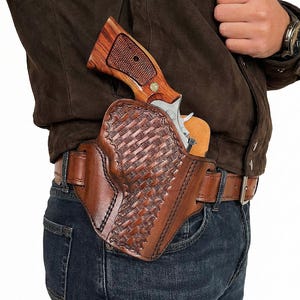 Può includere: Una fondina in pelle marrone con un motivo intrecciato, che contiene un revolver argentato con manico in legno. La fondina è attaccata a una cintura in pelle marrone indossata con jeans blu. L'impugnatura in legno del revolver è visibile.