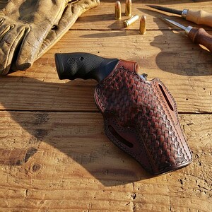 May include: A brown leather holster with a woven pattern holds a black revolver. A pair of tan leather work gloves, three bullets, and two wood-handled tools are also visible on a wooden surface. The holster is designed for a handgun.
