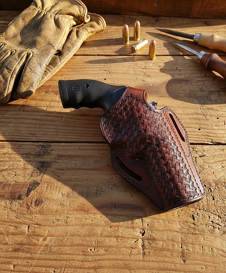 May include: A brown leather holster with a woven pattern holds a black revolver. A pair of tan leather work gloves, three bullets, and two wood-handled tools are also visible on a wooden surface. The holster is designed for a handgun.