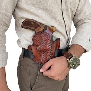May include: A brown leather holster with a basket-weave pattern, secured to a black belt. The holster holds a firearm with a dark wooden grip. The item is worn over brown trousers and a light beige button-down shirt, with a wristwatch visible.