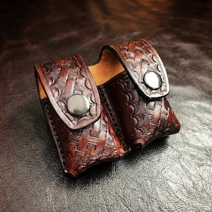Könnte beinhalten: Eine handgefertigte, braune Ledertasche mit einem dekorativen geprägten Muster. Die Tasche hat zwei Fächer und wird mit zwei silberfarbenen Druckknöpfen verschlossen. Das Leder hat eine satte, dunkelbraune Farbe.