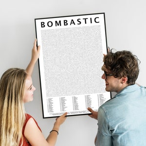May include: A black framed poster with the word "BOMBASTIC" at the top. The poster has a grid of small black dots and a list of words at the bottom. A man and woman are holding the poster.