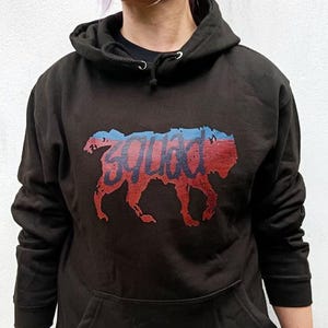 Puede incluir: Sudadera con capucha negra con bolsillo de canguro. La sudadera presenta un gráfico rojo y azul de una silueta de perro con la palabra "Squad" impresa. La sudadera tiene un cordón y un estilo casual.