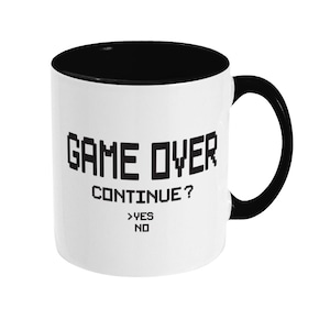 May include: White ceramic coffee mug with a black rim and handle. The mug has the text "GAME OVER CONTINUE? >YES NO" printed on it in black.