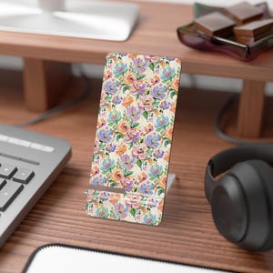 May include: A rectangular phone stand featuring a vibrant floral pattern with painted brushstrokes in shades of peach, lavender, blue, and green on a cream background. The stand has a clear acrylic support ledge at the bottom for holding a device.