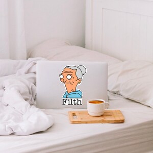 May include: A laptop computer with a sticker of a cartoon woman with grey hair and a blue hoodie. The sticker says "Filth".