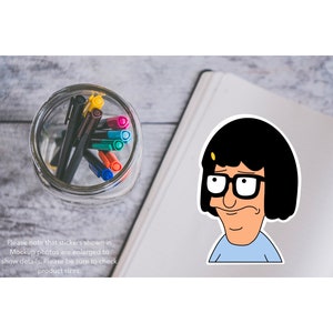Puede incluir: Una pegatina con un personaje de dibujos animados con cabello negro, gafas y una camisa azul. El personaje mira hacia la izquierda con una expresi&oacute;n ligeramente preocupada. La pegatina est&aacute; en una libreta blanca con un frasco de vidrio de bol&iacute;grafos de colores en el fondo.