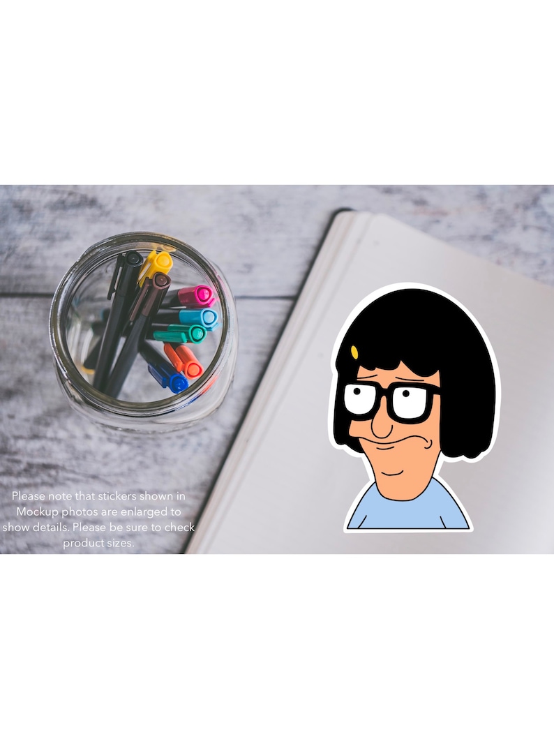 Puede incluir: Una pegatina con un personaje de dibujos animados con cabello negro, gafas y una camisa azul. El personaje mira hacia la izquierda con una expresi&oacute;n ligeramente preocupada. La pegatina est&aacute; en una libreta blanca con un frasco de vidrio de bol&iacute;grafos de colores en el fondo.