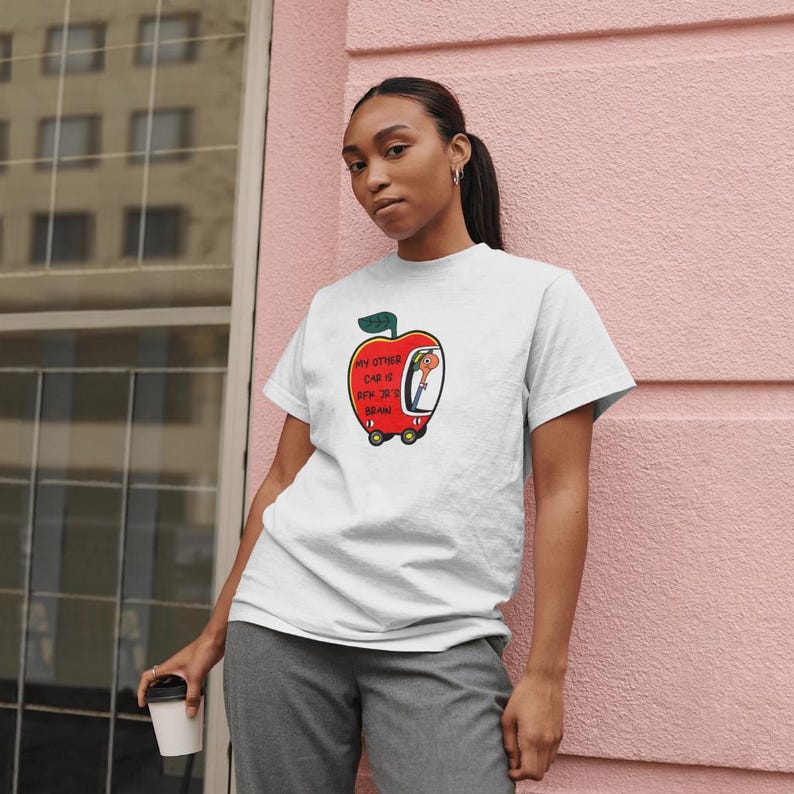 May include: White t-shirt with a red apple graphic. The apple has a face and is driving a car. The text on the shirt reads "My other car is a box truck brand".