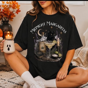 May include: A black t-shirt featuring a graphic print with the text Midnight Margaritas, a black cat, a bottle of tequila, a margarita glass with a lime wedge, a small bowl of salt, a book, a candle, and lavender sprigs under a crescent moon.