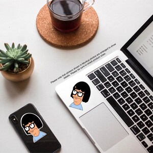 Puede incluir: Dos pegatinas con un personaje de dibujos animados con cabello negro, gafas y una camisa azul. Las pegatinas est&aacute;n en una computadora port&aacute;til y una funda de tel&eacute;fono negra.