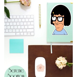 Puede incluir: Una pegatina con un personaje de dibujos animados con cabello negro, gafas y una camisa azul. La pegatina est&aacute; sobre un fondo verde claro. La pegatina est&aacute; sobre un escritorio con un teclado blanco, un bol&iacute;grafo dorado, una nota adhesiva azul claro y una alfombrilla de rat&oacute;n de cuero marr&oacute;n. Hay un rat&oacute;n blanco con una pegatina de rosa rosa en &eacute;l. Hay dos peon&iacute;as blancas en el escritorio.