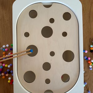 Könnte beinhalten: Ein Holz-Lernspielzeug mit weißem Rahmen und hellem Holzinnenraum mit unterschiedlich großen kreisförmigen Löchern. Eine Holzpinzette hält einen kleinen blauen Pompon, mit bunten Pompons, die um die Basis verstreut sind. Dieses Spielzeug ist für die Entwicklung der Feinmotorik konzipiert.