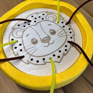 Könnte beinhalten: Ein gelbes und hölzernes Aktivitätsspielzeug mit Löwenmotiv. Das Spielzeug zeigt ein lächelndes Löwengesicht, das in einen Holzring geätzt ist, umgeben von einem gelben Rand. Braune und gelbe Pfeifenreiniger sind durch die Löcher gefädelt.
