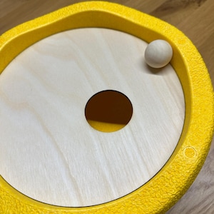 Könnte beinhalten: Ein Holzspielzeug mit einem gelben, strukturierten Rand und einer hellen Holzmitte. Eine kleine Holzkugel liegt auf der hellen Holzoberfläche. Ein Loch ist in die Mitte des Holzes geschnitten. Das Spielzeug ist für Kinder konzipiert.