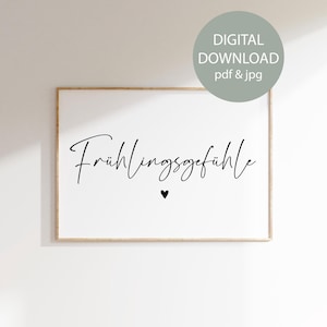 May include: A framed white print with the German phrase "Frühlingsgefühle" in elegant black script, meaning "Spring Feelings", and a small black heart. The print is in a light wood frame and includes a digital download option.