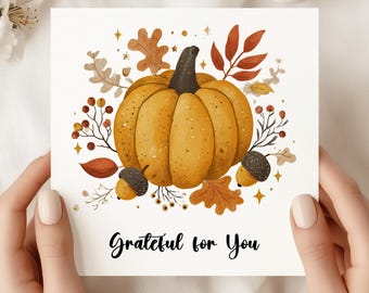 May include: A greeting card featuring a watercolor illustration of a pumpkin with a dark stem, surrounded by fall foliage and acorns. The card has the text "Grateful for You" in a handwritten style. The card is held by two hands.