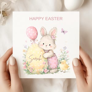 Peut inclure: Carte de Pâques carrée avec une illustration à l'aquarelle. Elle représente un lapin en robe rose, un œuf jaune avec le nom "Sarah", un ballon rose à pois et un poussin jaune. Le texte "HAPPY EASTER" est en haut.