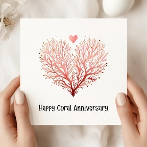 Könnte beinhalten: Quadratische Grußkarte mit einem korallenfarbenen herzförmigen Design und einem kleinen rosa Herz darüber. Der Text "Happy Coral Anniversary" ist in Schwarz. Die Karte wird von zwei Händen gehalten.