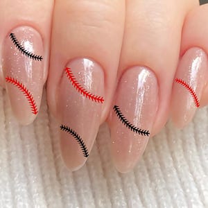 May include: Close-up of a hand with long, oval-shaped nails. The nails are painted with a light, shimmery nude polish and feature baseball-inspired stitching designs in red and black. The nails are set against a white, textured background.