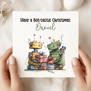May include: A square Christmas card featuring two cartoon robots, one yellow and one green, holding gifts. The card reads "Have a Bot-tastic Christmas Daniel" in black and green lettering.