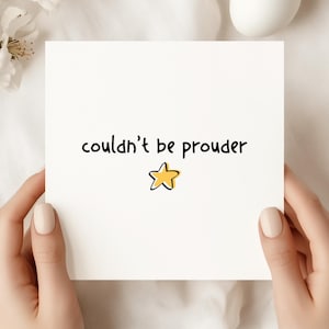 Könnte beinhalten: Eine weiße Karte mit dem Schriftzug "couldn't be prouder" in schwarzer Schrift und einer gelben Sternillustration. Die Karte wird von zwei Händen mit manikürten Nägeln gehalten. Der Hintergrund ist eine weiche, neutrale Farbe.