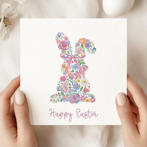 May include: A white card featuring a floral bunny design in shades of pink, purple, blue, and yellow. The bunny is composed of various flowers and has a pink bow. The words "Happy Easter" are written in cursive below the bunny.