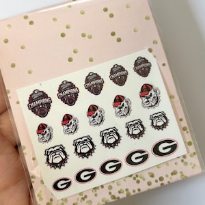May include: A sheet of nail decals featuring the University of Georgia Bulldogs logo, including the team mascot, the words "Champions", and the team's "G" logo.
