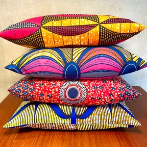 Op de afbeelding: Vier decoratieve kussens gestapeld op een houten oppervlak. Elk kussen heeft een uniek, levendig Afrikaans wasprintontwerp in verschillende kleuren, waaronder roze, geel, blauw en rood. De patronen omvatten geometrische vormen en bloemmotieven.