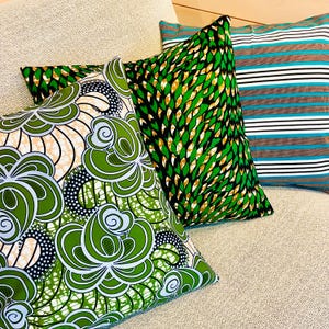 Puede incluir: Cuatro cojines decorativos con patrones vibrantes. Uno presenta un diseño floral verde, otro un patrón de hojas verdes y doradas, y un tercero muestra rayas azules y marrones. Los cojines están sobre una superficie de tono neutro.