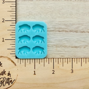 May include: A light blue silicone mold with six cavities shaped like buffalo silhouettes. The mold is approximately 1 inch wide and 1.5 inches tall.