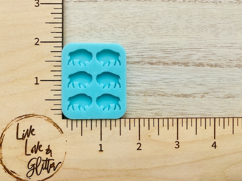 May include: A light blue silicone mold with six cavities shaped like buffalo silhouettes. The mold is approximately 1 inch wide and 1.5 inches tall.