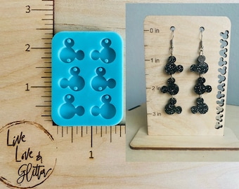 May include: A blue silicone mold with four Mickey Mouse head shapes. The mold is next to a ruler measuring 3 inches.  A pair of black glitter Mickey Mouse dangle earrings are shown on a wooden display stand with a ruler measuring 3 inches.