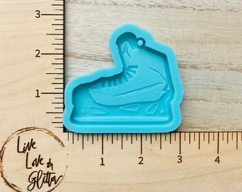 May include: A light blue silicone mold in the shape of an ice skate. The mold has a detailed design of the skate's laces, blade, and boot. The mold is on a wooden surface with a ruler and the text "Live Love & Glitter".