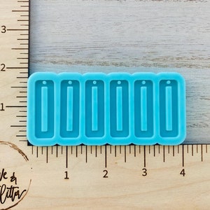 May include: A blue silicone mold with six rectangular cavities for making earrings or other jewelry. The mold is on a wooden surface with a ruler.