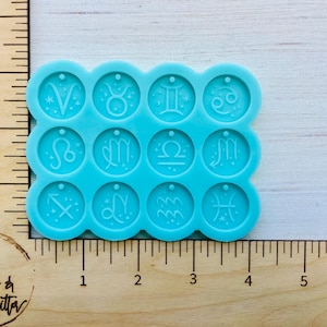 May include: A light blue silicone mold featuring twelve zodiac symbols. Each symbol is in a circular shape with a small hole at the top. The mold is placed on a wooden surface with a ruler and the text "Live Love & Glitter".