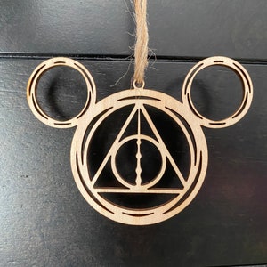 Könnte beinhalten: Holz-Anhänger in Form von Mickey-Maus-Ohren mit dem Symbol der Heiligtümer des Todes in der Mitte. Der Anhänger ist braun und hat einen rustikalen Look.