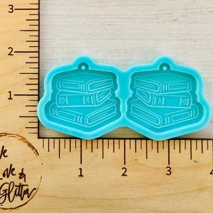 May include: A light blue silicone mold featuring two stacks of books. Each stack has three books with detailed spine and page edge designs. The mold is designed for crafting and has a small hole for hanging.